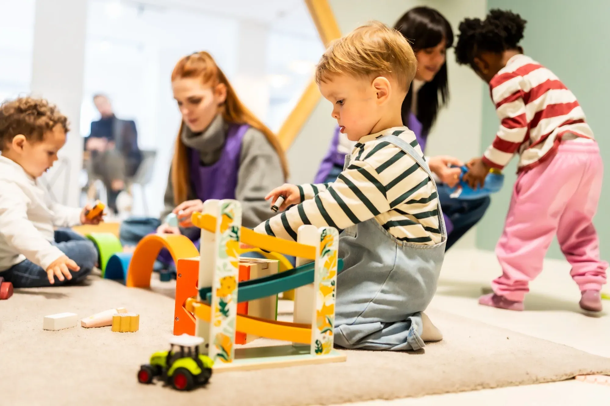 Toddlers playing with wooden toys on the floor of a Paris crèche while two carers supervise
